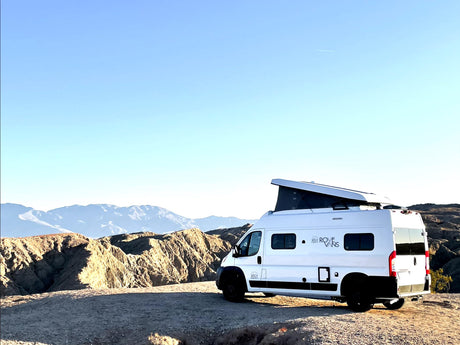 White RV rental van with pop-top roof atop a desert hill, overlooking rugged mountains under a clear sky.