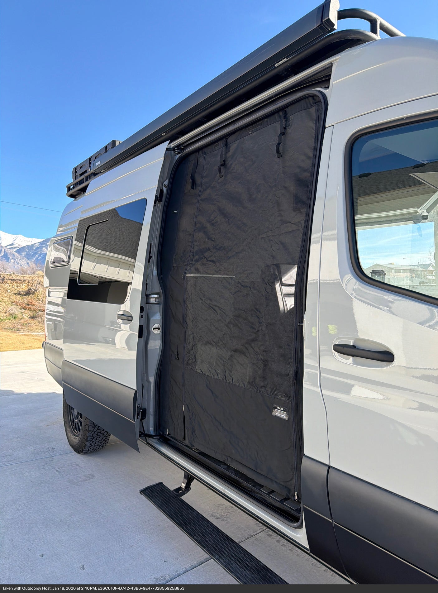 Light gray rv rental camper van with open sliding door, revealing a black bug screen & retractable step. Mountains in background.