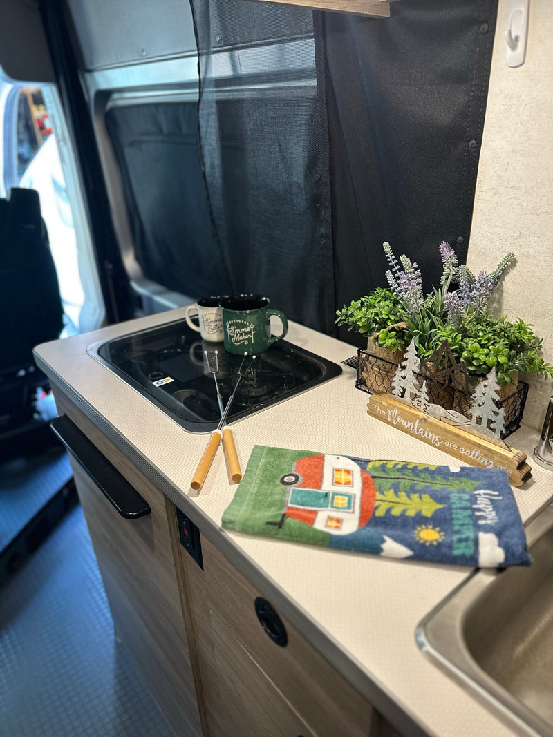 Experience a welcoming RV rental setup: kitchen counter with cooktop, mugs, roasting sticks, and a charming "Happy Camper" towel. — photo 13