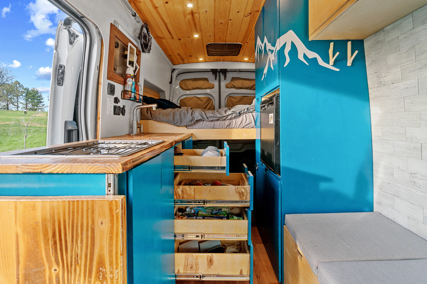 Explore this vibrant rv rental with a fully equipped kitchenette, pull-out storage, comfy bed, and mountain-themed blue interior.
