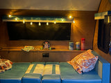 Relax and unwind in this cozy rv rental! Features warm string lights, a screen, and comfortable blue seating with patterned cushions.