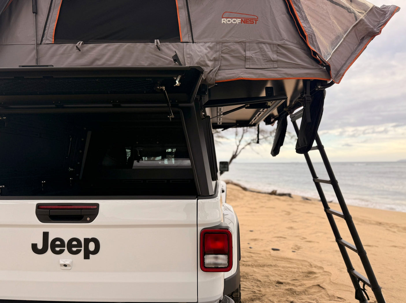 Explore with this rv rental: a white Jeep truck with an open Roofnest rooftop tent, parked right on a sandy beach by the ocean.
