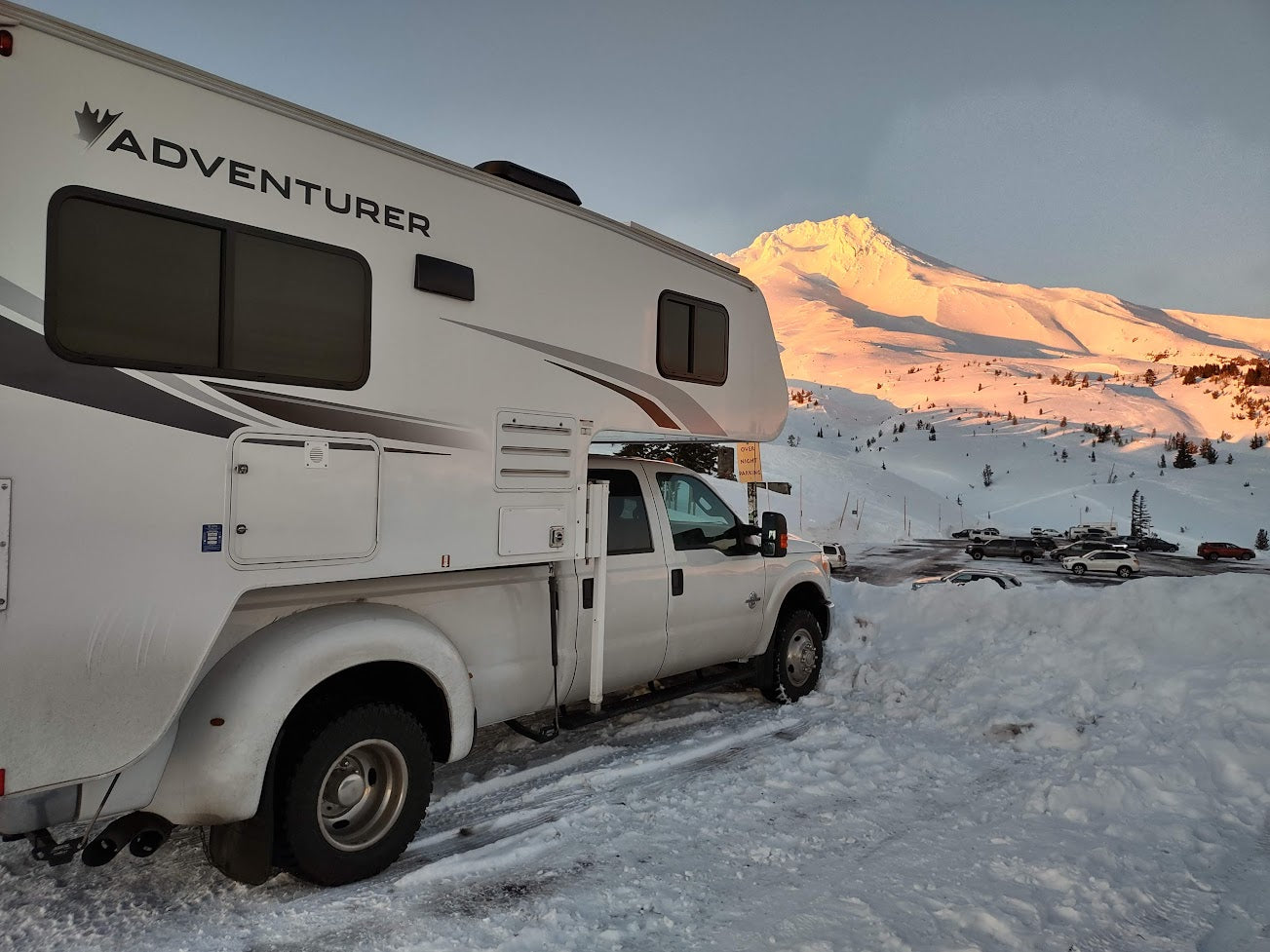 Adventurer RV rental truck camper parked in a snowy lot with a golden, sunlit mountain at dawn/dusk.