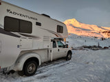 Adventurer RV rental truck camper parked in a snowy lot with a golden, sunlit mountain at dawn/dusk.