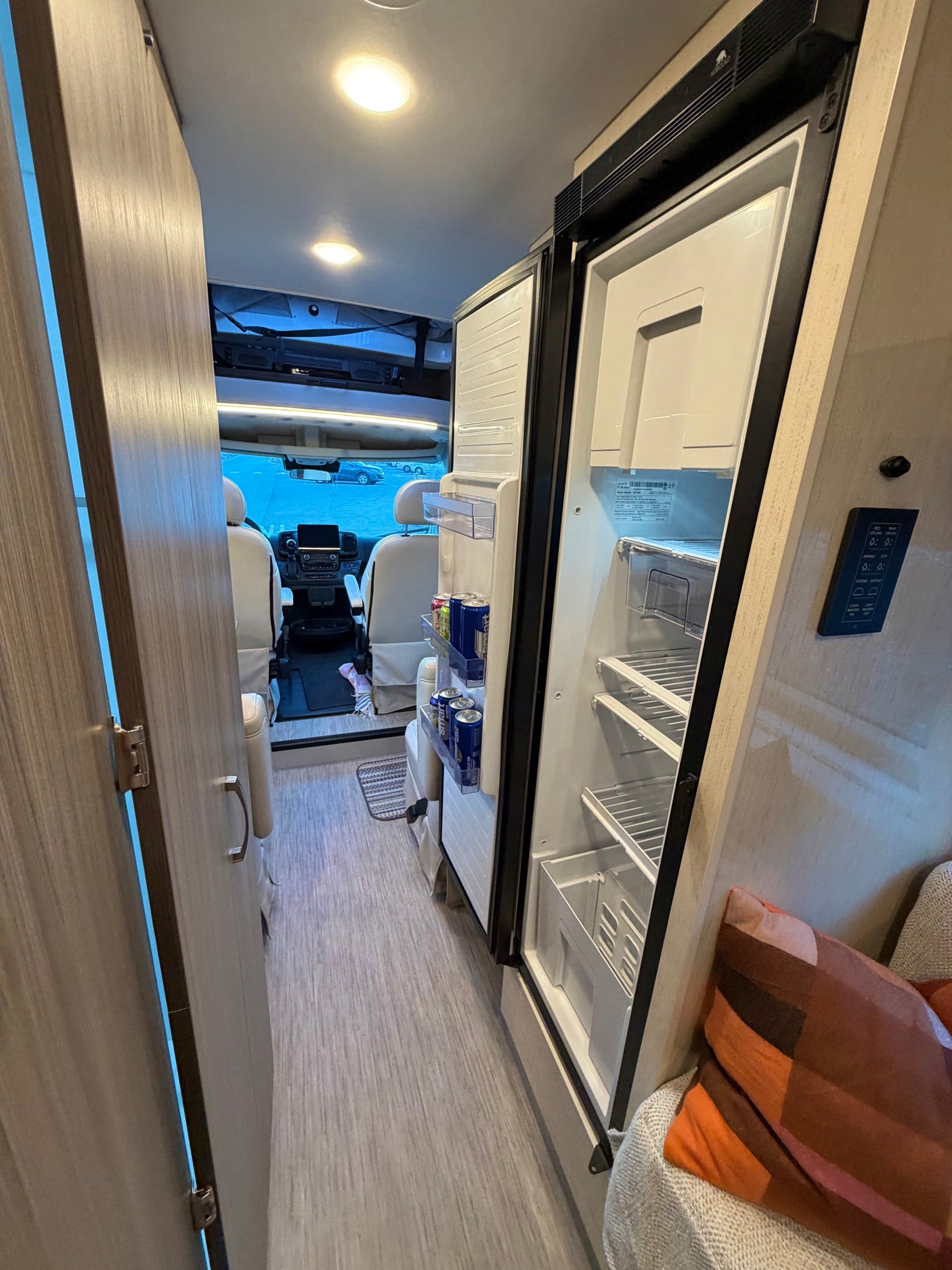 Inside an RV rental: open refrigerator showing drinks, sleek wood finishes, a patterned floor, and the cockpit view.