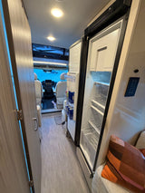 Inside an RV rental: open refrigerator showing drinks, sleek wood finishes, a patterned floor, and the cockpit view.