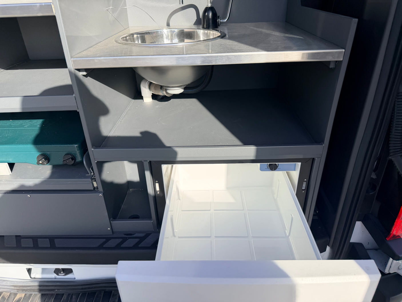 RV rental: Compact campervan kitchen features a stainless steel sink, portable stove, and a pull-out refrigerator drawer.
