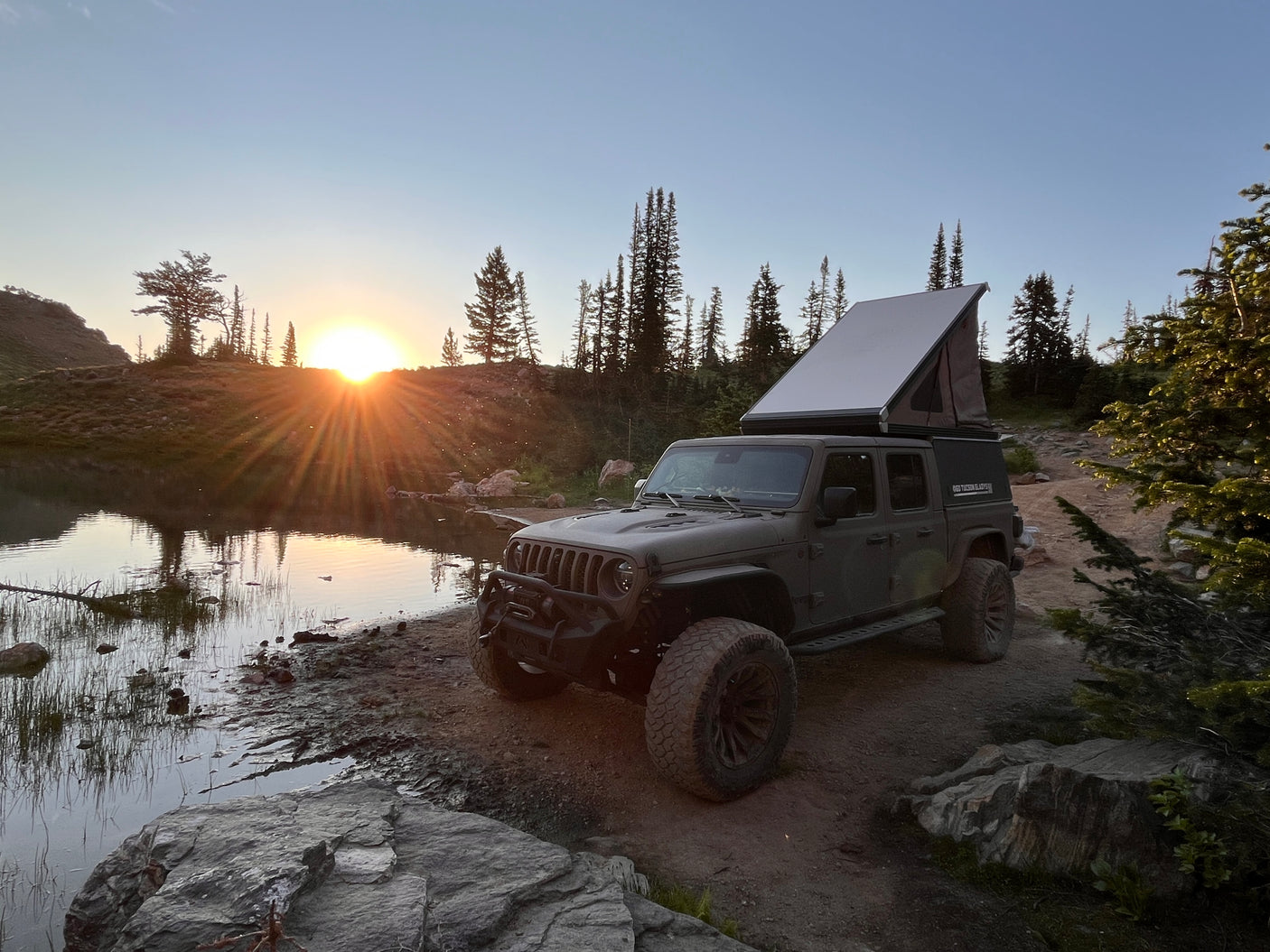 Adventure awaits! This rugged Jeep Gladiator with a pop-up camper, perfect for an RV rental, is parked by a scenic lake at sunset.