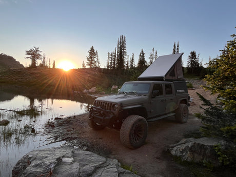 Adventure awaits! This rugged Jeep Gladiator with a pop-up camper, perfect for an RV rental, is parked by a scenic lake at sunset.