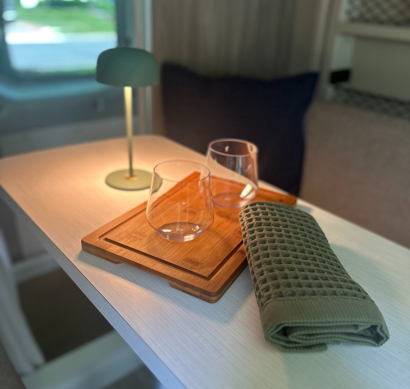 Cozy RV rental interior with a table, lamp, glasses, bamboo board, and green towel. Perfect for a relaxing trip.