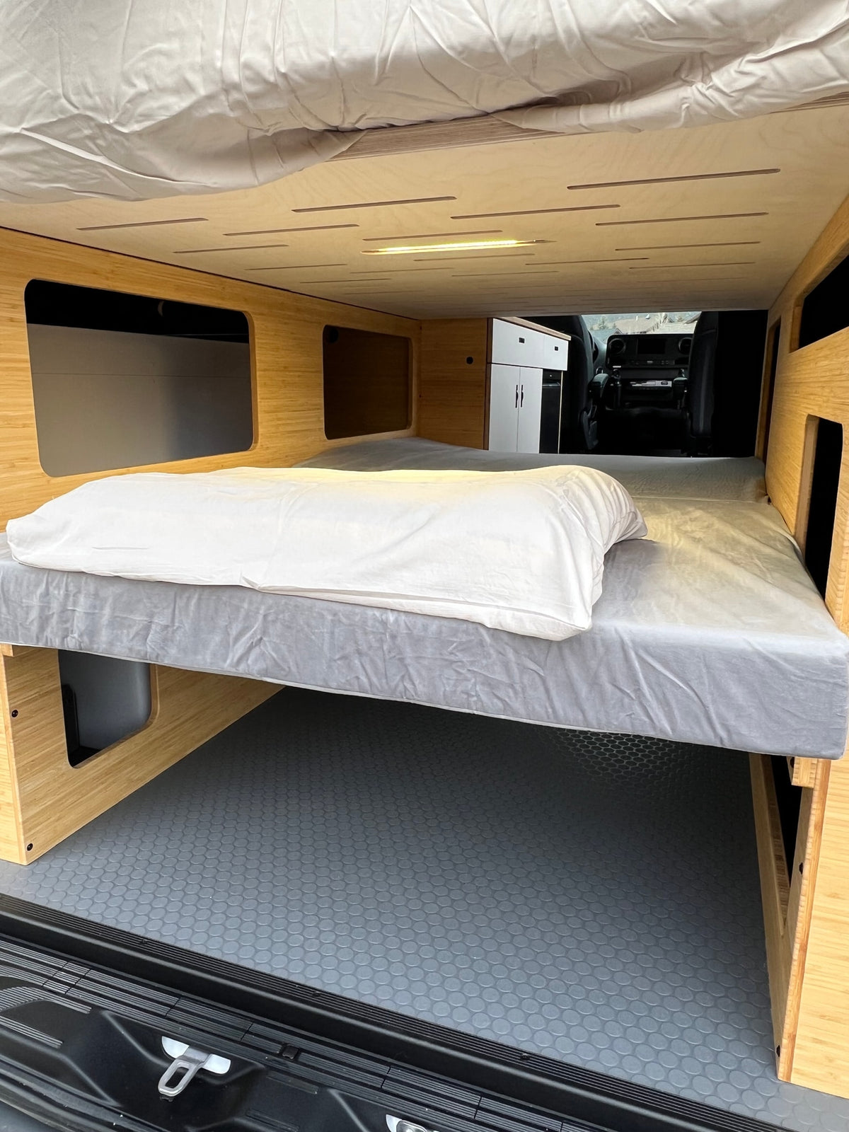 RV rental: Van interior with an elevated bed, white pillow, gray mattress, wooden storage, and a view into the front cab. — photo 20