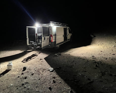 An RV rental camper van parked outdoors at night, with interior lights on and rear doors open, illuminating the surrounding rocky ground.