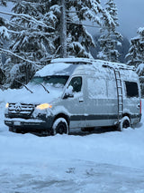 This snowy Mercedes Sprinter rv rental van, with headlights on, is nestled in a winter wonderland forest. Ready for cold-weather travel.