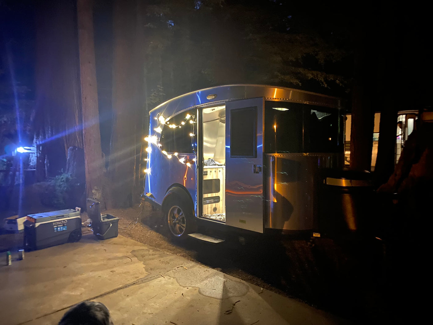 Night view of a sleek, silver rv rental trailer adorned with festive string lights, parked on a patio in a dark forest.