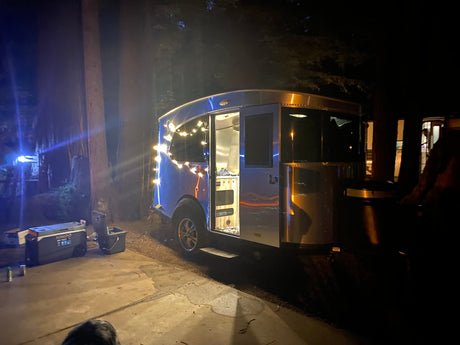 Night view of a sleek, silver rv rental trailer adorned with festive string lights, parked on a patio in a dark forest.