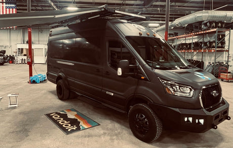 A custom dark grey Vandoit Ford Transit adventure van with off-road tires & roof rack, ready for RV rental, in a workshop.