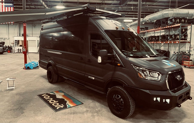 A custom dark grey Vandoit Ford Transit adventure van with off-road tires & roof rack, ready for RV rental, in a workshop.