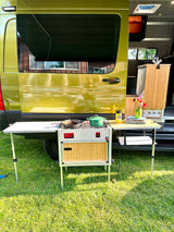 Outdoor kitchen set up next to a bright green RV rental, featuring a stove, pots, and a small dining table.