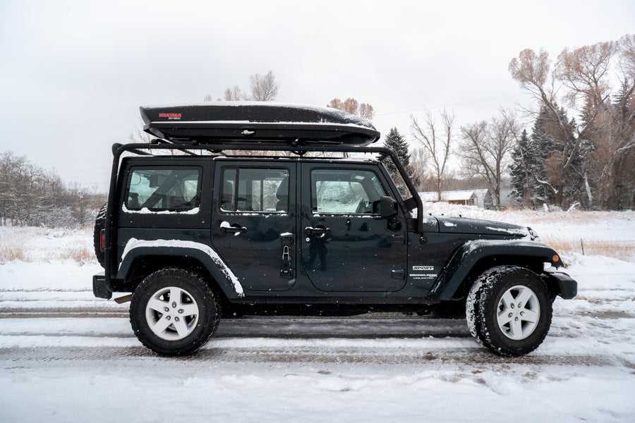 Dark Jeep Wrangler Unlimited with roof cargo box on a snow-covered road. Ideal winter adventure vehicle, perfect for an RV rental trip.