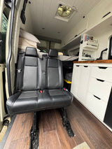 Interior of a custom RV rental featuring passenger seats, built-in kitchen area, and bed in the background.