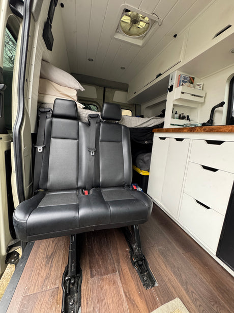 Interior of a custom RV rental featuring passenger seats, built-in kitchen area, and bed in the background.