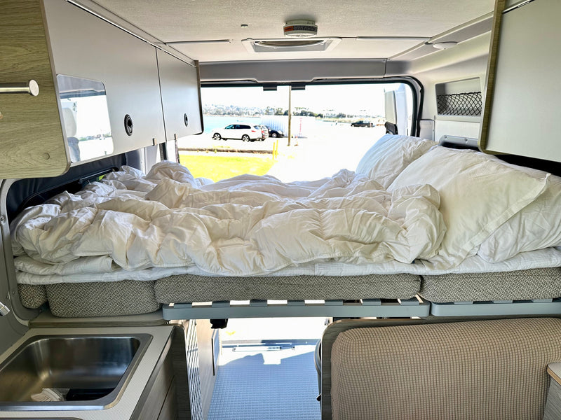 An RV rental camper van interior shows a cozy, raised bed with white linens, a sink, and scenic outdoor views. — photo 20