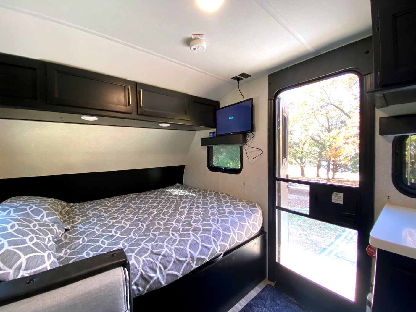 RV rental bedroom: A comfortable bed, overhead cabinets, wall-mounted TV, and an open door viewing autumn trees.
