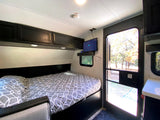 RV rental bedroom: A comfortable bed, overhead cabinets, wall-mounted TV, and an open door viewing autumn trees.