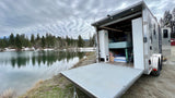 This RV rental offers lakeside camping! A travel trailer with its back open, revealing bunk beds by a calm lake.