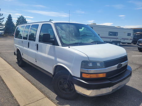 White Chevrolet Express passenger van in a sunny parking lot, with a large RV rental behind it.