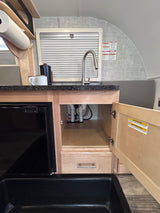 A compact RV rental kitchen features a black sink, faucet, small fridge, and open under-sink cabinet revealing plumbing.