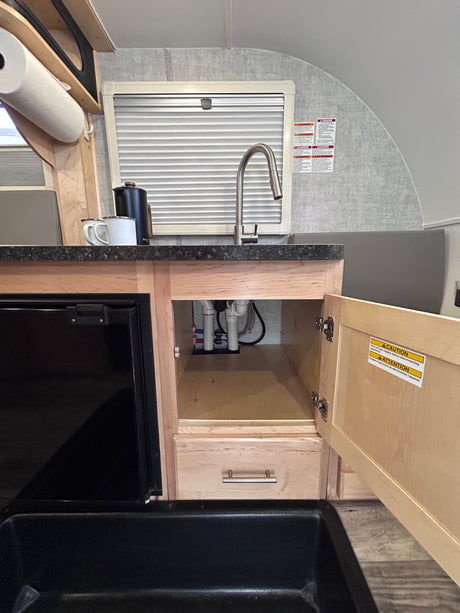 A compact RV rental kitchen features a black sink, faucet, small fridge, and open under-sink cabinet revealing plumbing.