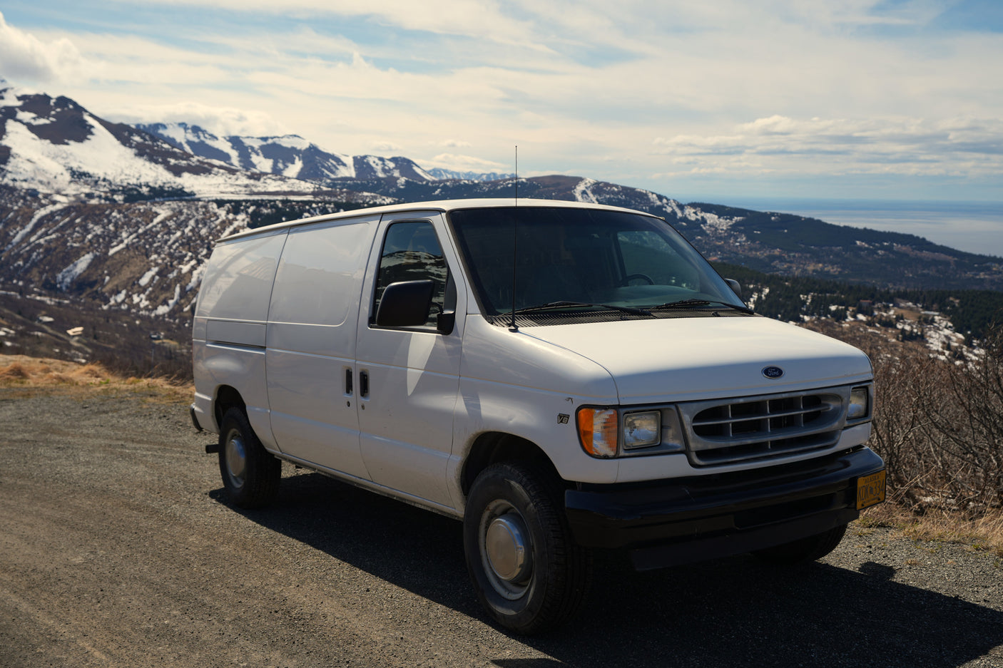White Ford van parked on a scenic mountain road overlooking snowy Alaskan peaks and a bay, ideal for an RV rental adventure.