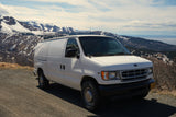 White Ford van parked on a scenic mountain road overlooking snowy Alaskan peaks and a bay, ideal for an RV rental adventure.