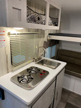 An RV rental interior featuring a compact kitchen with a two-burner stove, sink, cabinets, and a dinette/bunk area.
