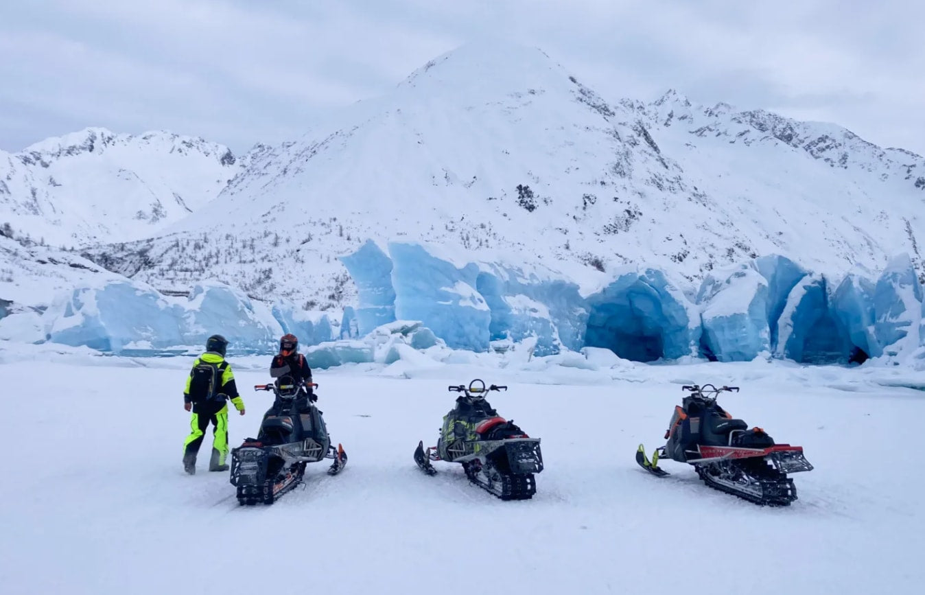 Experience glacier snowmobiling: two people & three snowmobiles on ice with blue glacier caves & snowy mountains. Plan your RV rental winter adventure!