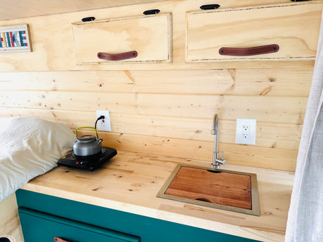 Cozy RV rental interior with natural wood walls, a small kitchen featuring a hot plate, sink, and overhead storage. Perfect for travel.