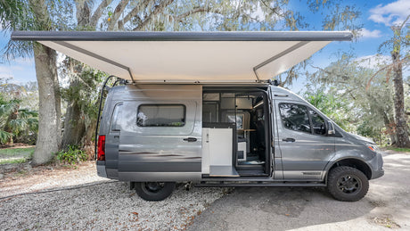 Grey Winnebago camper van for rv rental, parked in nature with awning extended & side door open.