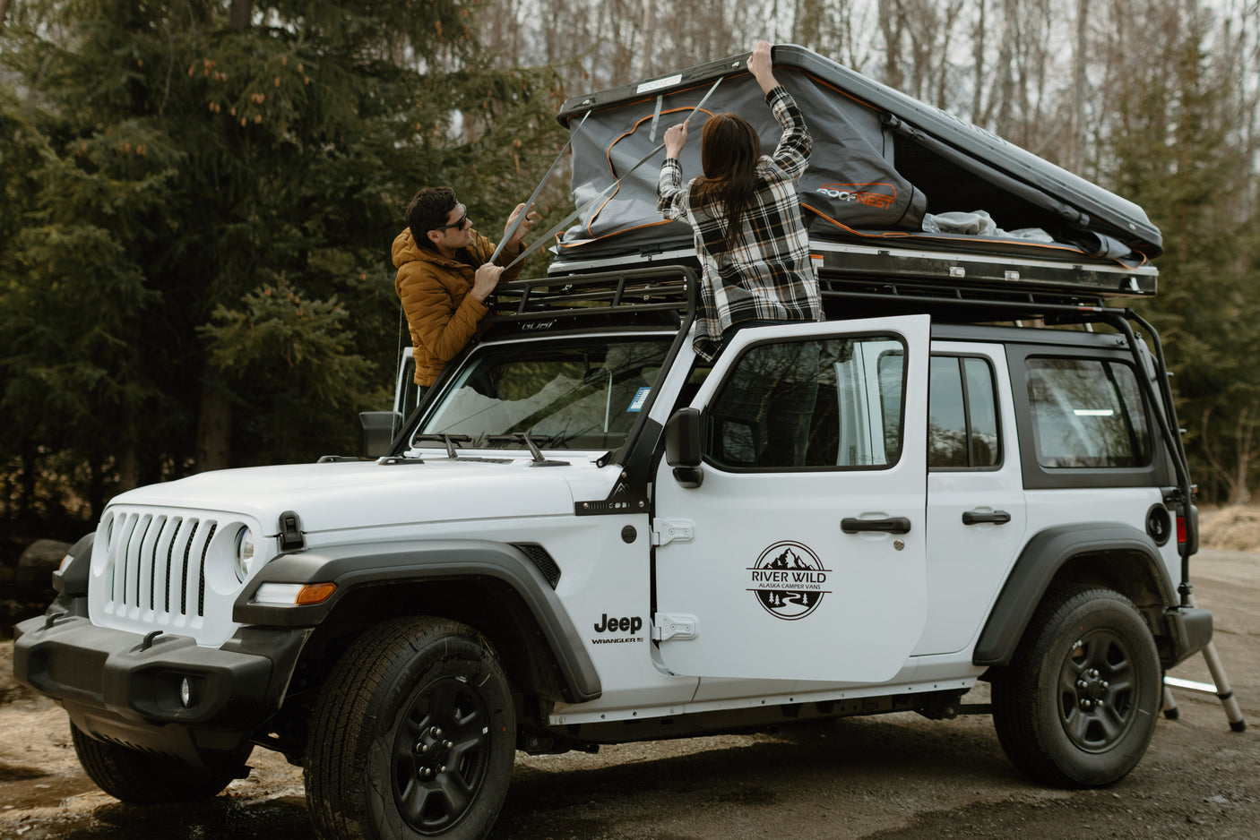 A white Jeep Wrangler RV rental with a rooftop tent being set up by two people, ready for a wilderness adventure.