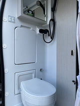 RV rental compact wet bath with a white toilet, shower, mirror, and light/fan controls on the wall.