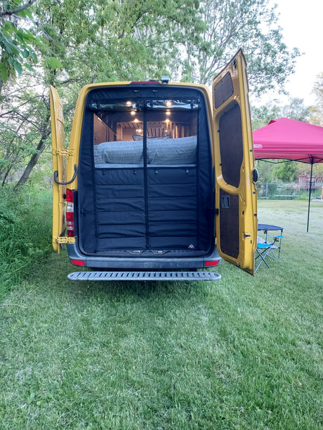 Yellow camper van (RV rental) with open rear doors, revealing a cozy interior bed with lights, parked on green grass.