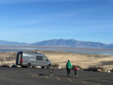 Enjoy an RV rental road trip! Family, dog, and a camper van by a scenic lake and mountains under a blue sky.
