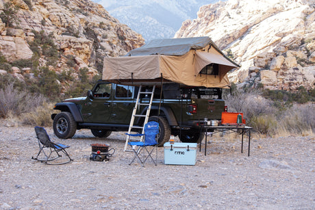 Experience off-grid camping with an RV rental! Jeep Gladiator, rooftop tent, cooler, chairs, and stove in a desert landscape.