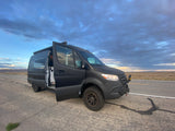 Adventure-ready matte black Sprinter van RV rental parked on a desert road with doors open under a cloudy sky.