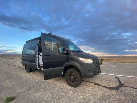 Adventure-ready matte black Sprinter van RV rental parked on a desert road with doors open under a cloudy sky.