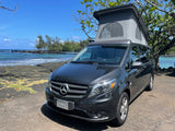 Dark gray Mercedes-Benz pop-top camper van rv rental parked on a sunny rocky coast by blue ocean waves.