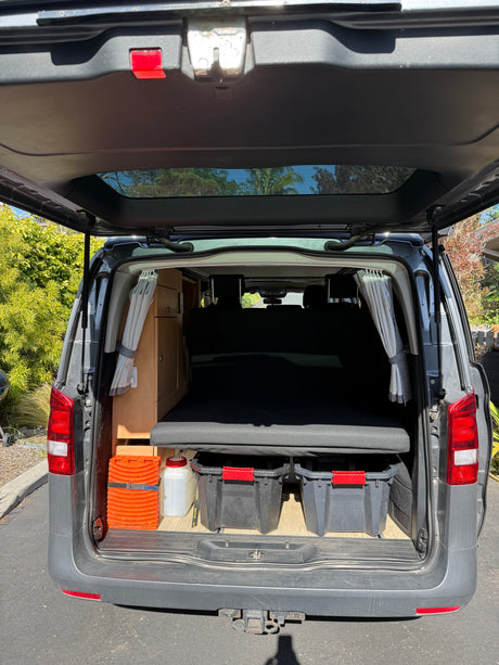 Open tailgate view of an RV rental camper van with a cozy bed, handy storage bins, and wood cabinets.