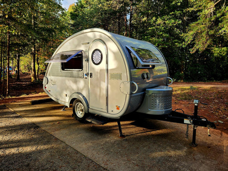 A sleek, silver teardrop RV rental sits in a sun-dappled forest campsite, perfect for a nature getaway.