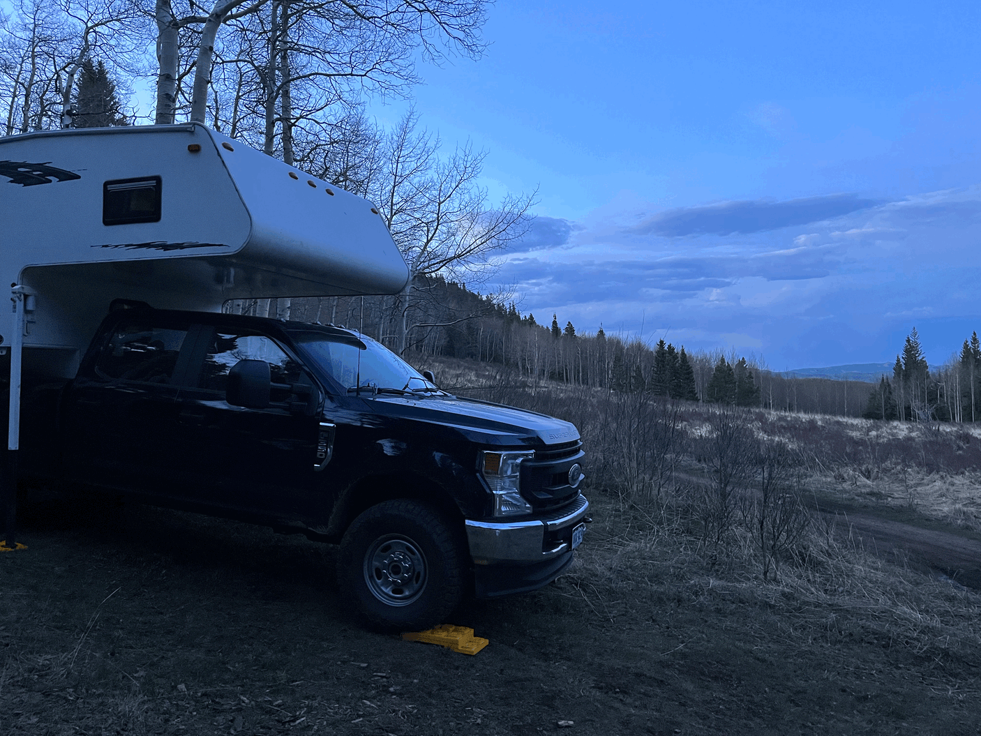 Black truck with white truck camper parked on a dirt path under a vast blue sky in a bare tree landscape. Ideal RV rental escape!