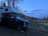 Black truck with white truck camper parked on a dirt path under a vast blue sky in a bare tree landscape. Ideal RV rental escape!